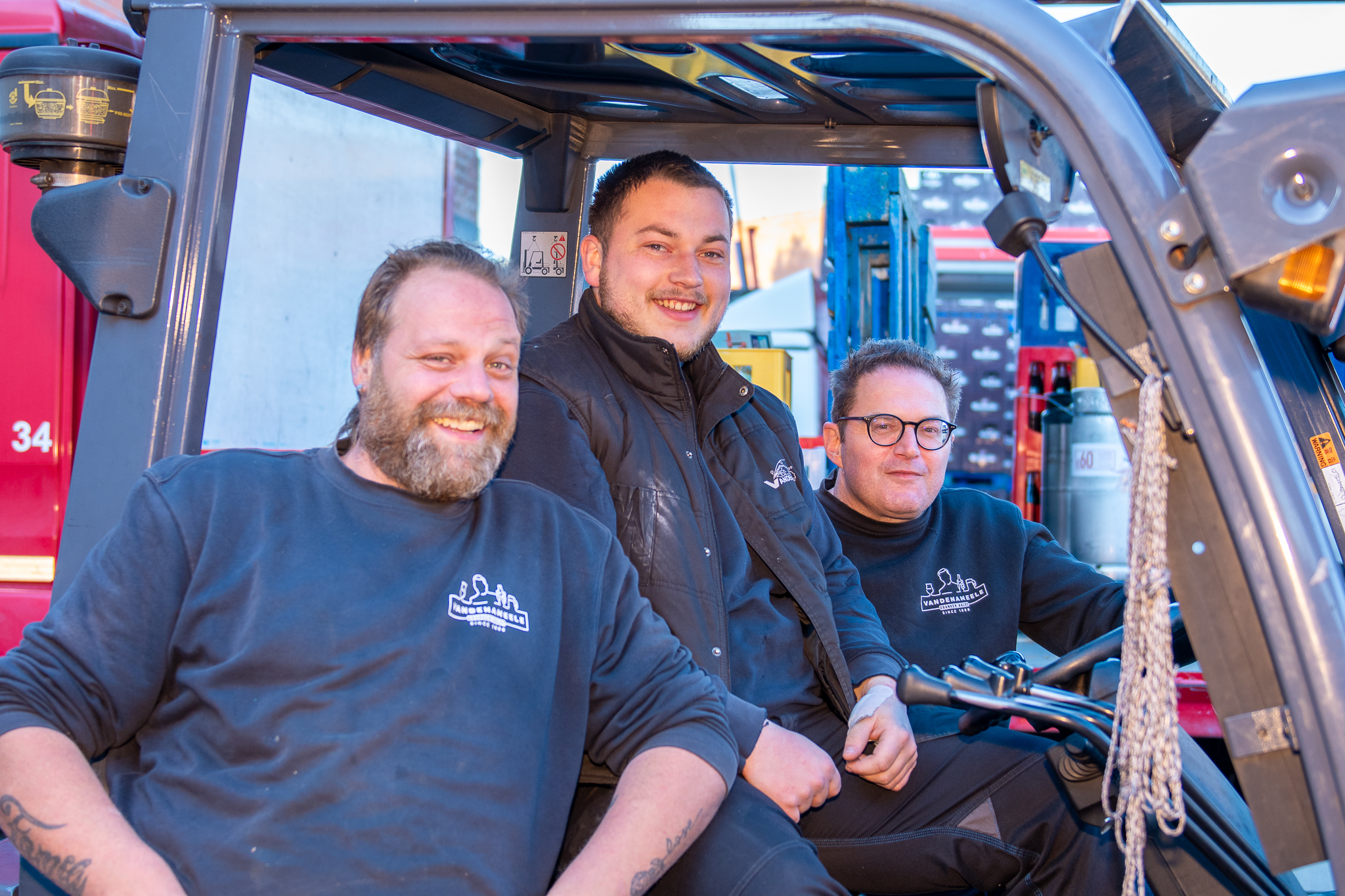 Drie enthousiaste medewerkers van Dranken Vandenameele poseren samen op een heftruck in het distributiecentrum.